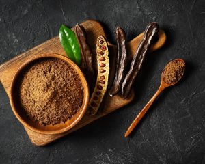Carobpulver in einer Holzschale, Johannisbrotschoten und ein Holzlöffel auf dunklem Untergrund.