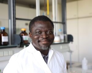 Richard Atuna im weißen Laborkittel vor Glasgeräten und Chemikalienflaschen im Labor für Lebensmittelanalytik.