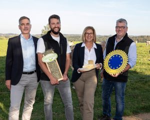 Vier Personen präsentieren Heu, Käse und das EU-Gütesiegel garantiert traditionelle Spezialität vor einer Kuhweide