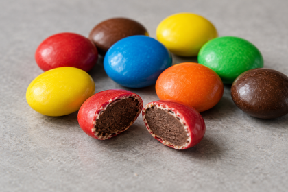Bunte Schokoladendragees auf hellem Untergrund, vorne ein aufgebrochenes rotes Dragee mit sichtbarem Schokoladenkern.