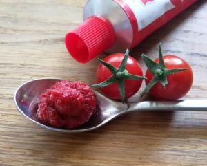 In einem Löffel befindet sich Tomatenmark; daneben eine Tube mit Tomatenmark und zwei kleine Cocktailtomaten.